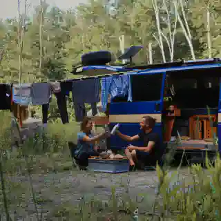 Jo and Georg eating infront of their T4 Campervan. They sit in camping chairs, next to hanging laundry. The Starlink Dish is mounted on the vans rooftop terrace.