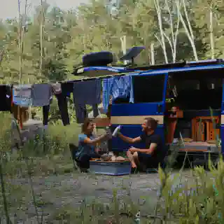 Jo und Georg sitzen auf Campingstühlen vor ihrem T4 Campervan. Man sieht Wäsche auf der Leine und die Starlink Dish auf der Dachterasse.