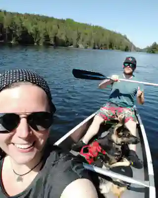 Jo, Georg und Momo im Kanu. Jo sitzt vorn und nimmt das Selfie auf, Georg hinten und hält ein Paddel in der Hand. Momo liegt in der Mitte des Kanus.