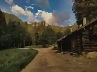 A wooden alpine hut on the right side. The path winds past the hut into  the forest behind it. Further back, you can see the mountains