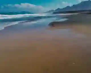 Auf der linken Seite ist das Meer zu sehen rechts ein endloser Sandstrand. Im Hintergrund sind Berge zu sehen. Ganz klein kann man Jo, Nelly und Momo am Strand sehen 