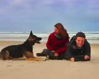 Standstrand mit Meer und Wellen. Momo, Jo und Georg im Vordergrund. Momo und Jo schauen sich an während Georg in die Kamera blickt. 