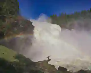 Der Wasserfall mit einem Regenbogen. Auf einem Stein vor dem Wasserfall sitzt Jo.