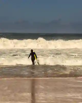 Jo trägt das Surfboard in das Meer, hinter Ihr sieht man Wellen