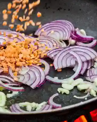 Eine Pfanne mit Öl und Zwiebeln. Rote Linsen fallen von oben in die Pfanne.