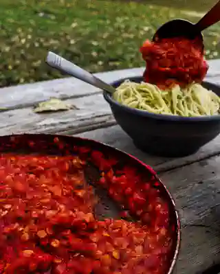 Eine Pfanne mit roter Linsenbolognese auf einem Tisch stehend. Im Hintergrund steht eine Schale mit Spagetti auf die gerade eine Kelle mit Bolognese gegossen wird