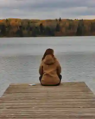 Jo sitzt auf einem Steg an einem See. Am anderen Ufer ein herbstlicher Wald.