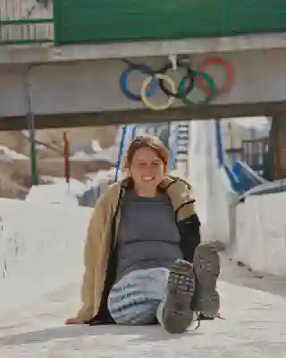 Auf der Bobbahn. Jo sitzt auf der Bobbahn ohne Eis. Im Hintergrund sind die 5 Olympischen Ringe zu sehen.